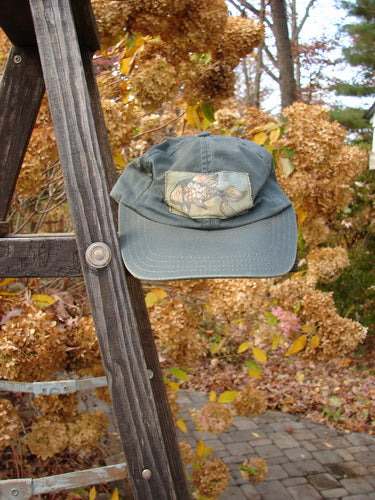 A faded green 1999 Patched Men's Baseball Cap Goldfish Blue Fish Logo Hunter Teal OSFA hangs on a weathered wooden stepladder amidst an autumnal garden, showcasing its distinct fish patch and vintage charm.