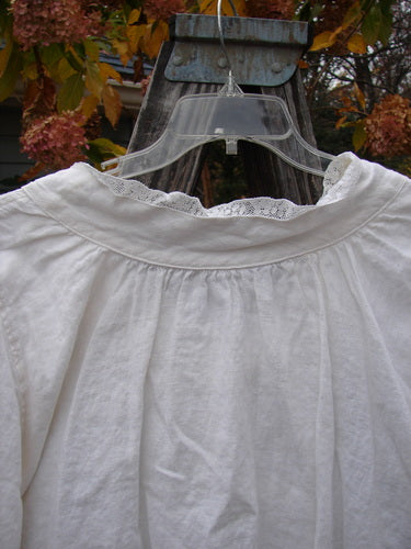 Magnolia Pearl Homespun Linen Lace Button Bib Top displayed on a wooden stand, featuring lace accents, cloth-covered buttons, and a unique silhouette with billowed sleeves and varying hemline.