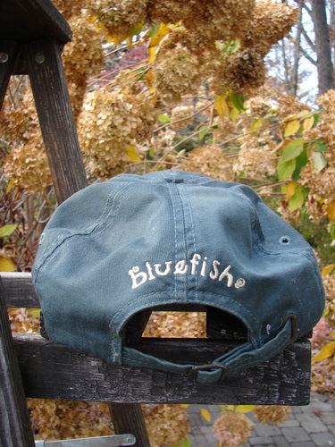 1999 Patched Men's Baseball Cap Goldfish Blue Fish Logo Hunter Teal OSFA, features a goldfish patch, adjustable rear tan strap, and slight fade on brim. Embroidered 'Bluefish' on back, resting on wooden surface.