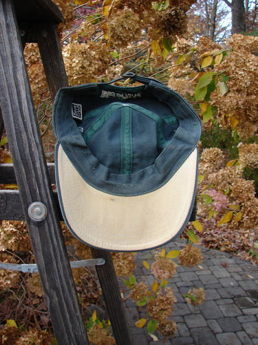 1999 Patched Men's Baseball Cap Goldfish Blue Fish Logo Hunter Teal OSFA draped over a wooden ladder rung, showcasing its tan brim, goldfish patch, and adjustable rear, with autumn elements subtly behind.