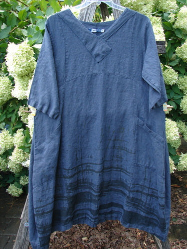 Barclay Linen Cross Over Incense Dress in structural cliff deep navy, size 0, made from textured linen with a relaxed V-neck crossover front and loose short sleeves. The dress hangs outdoors against leafy greenery, showing a softly draped silhouette and darker horizontal banding near the hem.