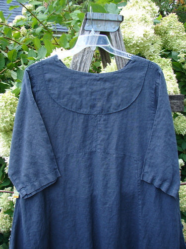 Barclay Linen Cross Over Incense Dress in structural cliff deep navy, size 0, hangs from a clear hanger outdoors. The linen fabric has a softly textured finish, a rounded crossover neckline, and short sleeves, with the deep blue dress falling in a relaxed, straight silhouette.