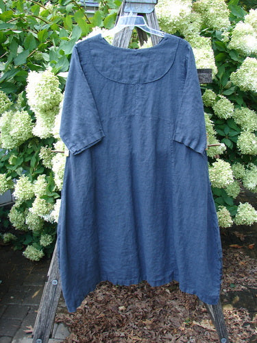 Barclay Linen Cross Over Incense Dress Structural Cliff in deep navy, size 0, made from textured linen with a relaxed A-line shape and short sleeves. The dress hangs on a clear hanger against leafy white hydrangeas, with soft natural light emphasizing its matte drape.
