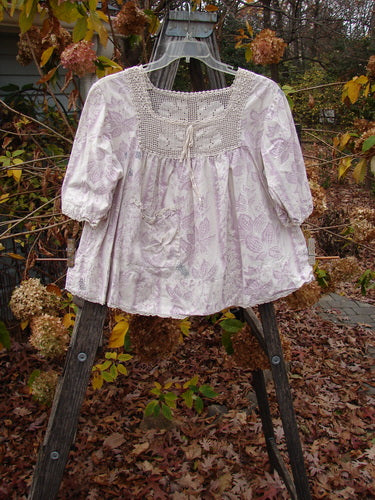 Magnolia Pearl Homespun Linen Crochet Bunny Blouse Swirl Lavender OSFA displayed on a swing, highlighting its crochet squared neckline, lace-edged sleeves, and front lace-topped pocket.