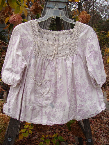 Magnolia Pearl Homespun Linen Crochet Bunny Blouse Swirl Lavender OSFA displayed on a rack, featuring a crochet squared neckline, three-quarter billowed sleeves, and a lace-edged hem, exemplifying vintage-inspired craftsmanship.