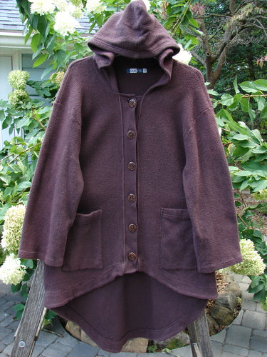 Barclay Celtic Moss Hooded 2 Pocket Jacket Unpainted Brum Size 0 displayed on a fence, showcasing its cozy hood, oversized buttons, and unique upward scooped front hemline.