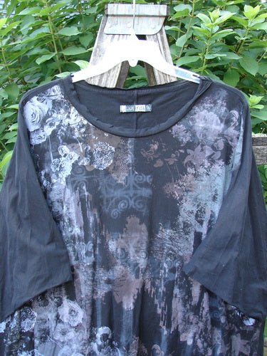 Barclay Raw Edge Flutter Collar High Low Top Dogwood Shadow Black OSFA, featuring a floral pattern, flutter neckline, short dolman sleeves, and a high-low hemline, displayed on a wooden surface.