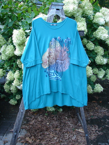 Barclay Banded Sleeved Art Top Wild Flower Vase in aqua, Size 3, a loose T-shirt with wide banded sleeves and a flowing hem. The front shows a vase-and-wild-flower graphic in soft blue and pink tones, hanging outdoors against white blossoms and greenery.