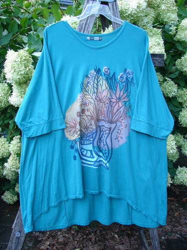 Barclay Banded Sleeved Art Top Wild Flower Vase in aqua, size 3, with wide banded sleeves and a loose drape. The soft jersey top features a centered wildflower vase graphic in layered blue, cream, and lavender tones, hanging on a clear hanger against leafy greenery.
