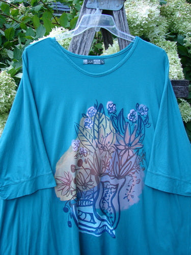 Barclay Banded Sleeved Art Top Wild Flower Vase in aqua, Size 3, with a loose banded-sleeve silhouette and a large floral vase graphic across the front. The soft knit fabric hangs from a clear hanger, and the vivid blue-green color stands out against the leafy outdoor background.