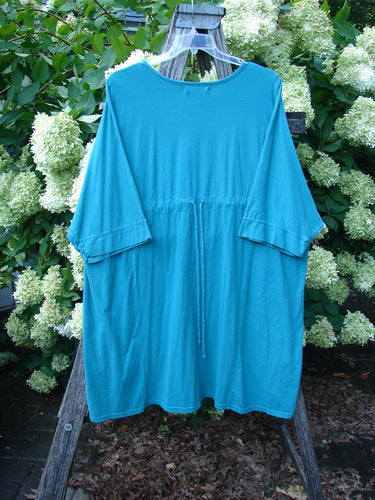 Barclay Banded Sleeved Art Top Wild Flower Vase in aqua, size 3, shown as a loose, short-sleeved top with a wide banded hem and soft drape. The smooth fabric hangs from a wooden hanger, set against dense white flowering foliage and dark ground cover.