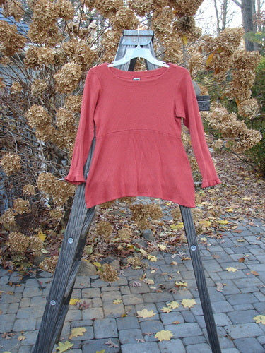 2000 Wool Pointelle Anais Top Unpainted Bittersweet Tiny Size 1: A red shirt on a wooden rack, featuring sweet lettuce edging, an empire waist seam, and a tailored waistline.