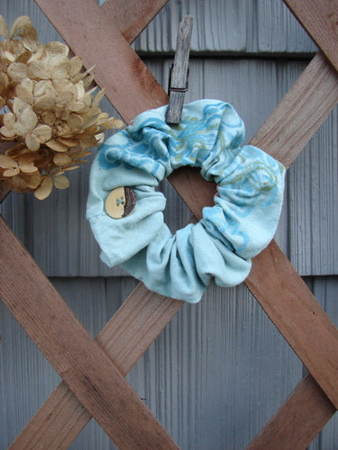 Barclay PMU Organic Cotton Button Hair Scrunchie Abstract Green Pink Medium displayed on a wooden fence, highlighting its ruffled fabric and elastic band, celebrating Jennifer Barclay’s sustainable vintage design collection.