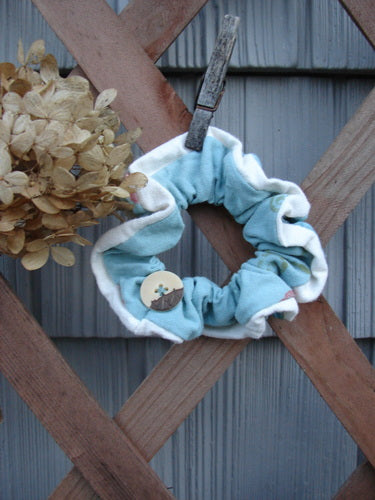 Barclay PMU Organic Cotton Button Hair Scrunchie Abstract Aqua Cream Medium displayed on a wooden fence, showcasing its ruffled fabric and button detail, embodying Bluefishfinder.com’s vintage, sustainable fashion ethos.