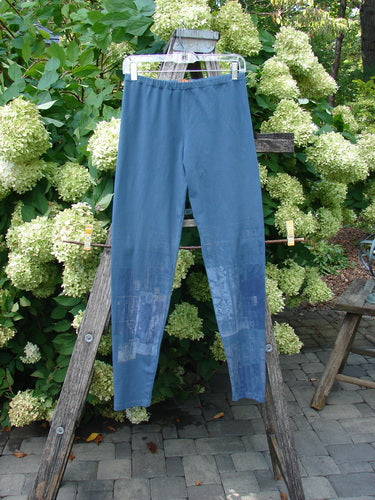 Barclay Cotton Lycra Legging Graphic Garden Echo Blue Size 1 in muted blue, with a slim stretch silhouette and subtle graphic patterning fading down the legs. The leggings hang from a wooden ladder outdoors, against dense green hydrangea blooms and gray stone paving.