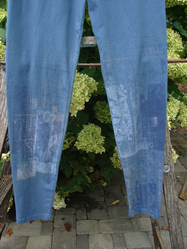 Barclay Cotton Lycra Legging Graphic Garden Echo Blue Size 1 in soft blue cotton-lycra, shown as long, slim leggings with a subtle graphic print and slightly faded panels. The fabric hangs straight in front of dense green foliage, creating a cool, muted contrast.
