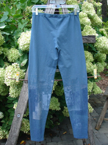 Barclay Cotton Lycra Legging Graphic Garden Echo Blue in size 1, shown as slim blue leggings with a smooth stretch finish and a subtle graphic print concentrated on the lower legs. They hang front-facing on a hanger outdoors, with a soft matte look against leafy greenery and pale blossoms.