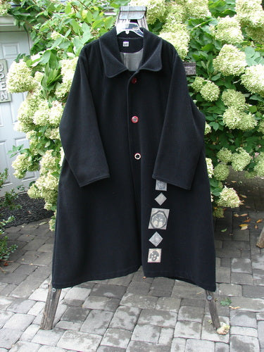 1999 Patched Woven Wool Overcoat in jet black, size 2, with a loose, long silhouette, broad notched collar, and oversized front buttons. The woven wool fabric hangs in a matte drape, while patch details line the lower front, and the coat is shown open on a stand against leafy greenery.