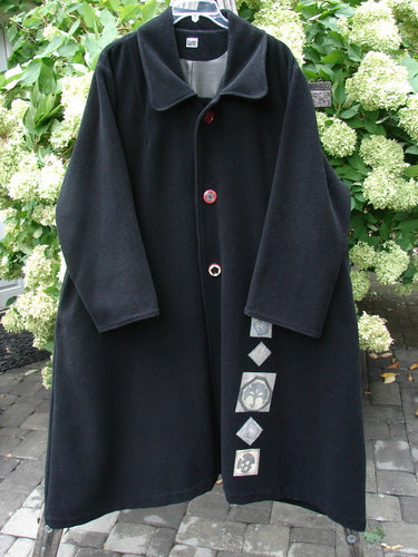 1999 Patched Woven Wool Overcoat in jet black, size 2, with a broad notched collar, oversized drape, and long sleeves. The wool surface looks dense and matte, while a vertical row of patch details and red-edged buttons adds contrast against the dark coat.