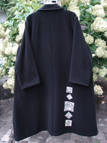 1999 Patched Woven Wool Overcoat in jet black, size 2, with a long, boxy silhouette, wide collar, and straight sleeves. The dense wool surface falls in a clean dark drape, while a vertical row of patched labels adds a subtle graphic detail near the lower back.