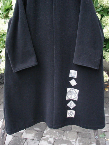 1999 Patched Woven Wool Overcoat in jet black, size 2, with a long straight silhouette, wide sleeves, and a softly textured wool surface. A vertical row of small square patch appliqués runs near the lower front, while the coat hangs centered against leafy greenery and stone paving.