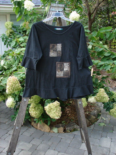 Barclay PMU Patched Flutter Top Wind Flower Black Size 2 displayed on a wooden ladder, showcasing the lightweight organic cotton fabric, rounded hem, three-quarter flutter sleeves, and wind flower theme patches.