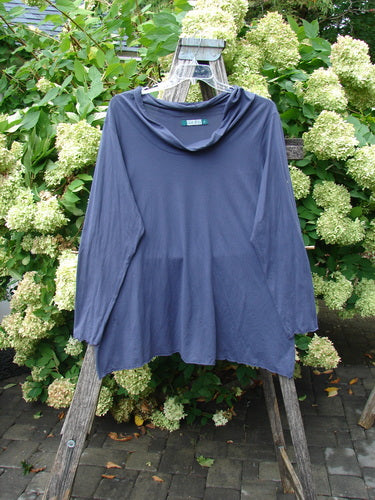 Barclay Batiste long-sleeved cowl neck layering top in unpainted royal orchid, size 2, shown draped on a weathered wooden ladder. The soft, fluid fabric falls in a relaxed silhouette with a loose cowl neckline and long sleeves, set against dense green foliage and pale blossoms.