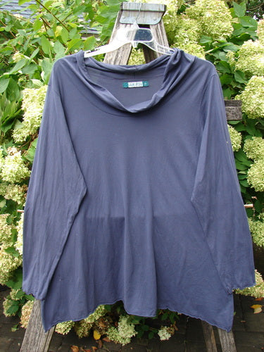 Barclay Batiste long-sleeved cowl neck layering top in unpainted royal orchid, size 2, shown in a soft drape with a loose neckline and gently flared hem. The smooth, matte fabric hangs from a hanger against leafy greenery, creating a deep blue-purple silhouette.