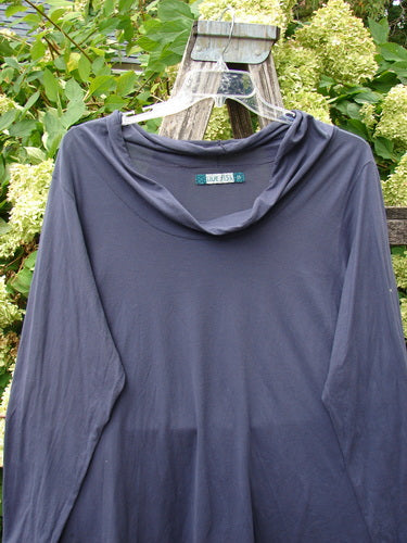 Barclay Batiste long-sleeved cowl neck layering top in unpainted royal orchid, size 2, shown in a soft drape with a loose cowl neckline and long sleeves. The muted purple knit hangs from a clear hanger against leafy greenery.