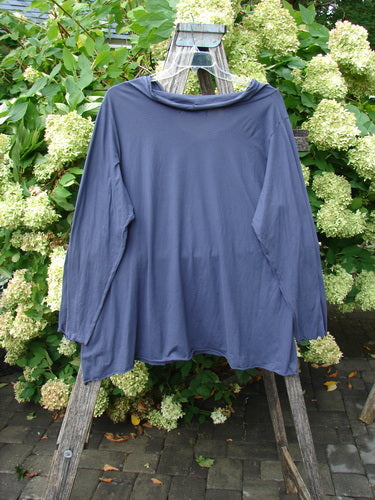 Barclay Batiste long-sleeved cowl neck layering top in unpainted royal orchid, size 2, shown in a soft blue-purple knit with a draped neckline and relaxed silhouette. It hangs from a weathered wooden ladder against dense green foliage and pale hydrangea blooms, with diffused daylight softening the folds.
