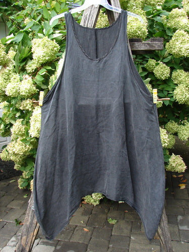 Barclay Hemp Silk Pinafore in unpainted smoke, size 2, with a loose sleeveless A-line shape and deep curved neckline. The dark gray hemp-silk fabric hangs softly from a hanger, its airy drape set against leafy greenery and weathered wood.