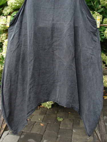 Barclay Hemp Silk Pinafore in unpainted smoke, size 2, with a loose, draped silhouette and soft hemp-silk texture. The dark gray garment hangs in a broad A-line shape, its hem curving into a central opening, against leafy greenery and stone paving.