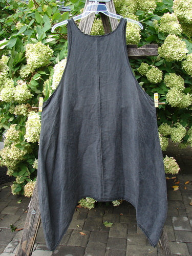 Barclay Hemp Silk Pinafore in unpainted smoke, size 2, with a loose A-line silhouette, narrow shoulder straps, and a softly textured drape. The dark charcoal fabric hangs from a hanger outdoors, framed by leafy greenery and weathered wood.