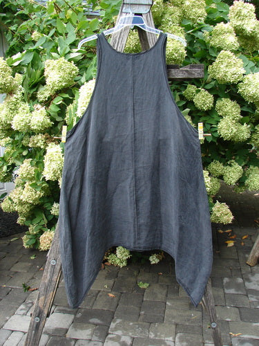 Barclay Hemp Silk Pinafore in unpainted smoke, size 2, hangs from a clear hanger with a loose, A-line silhouette and deep arm openings. The textured hemp-silk fabric falls in a soft charcoal drape, set against leafy green hydrangeas and weathered wood.