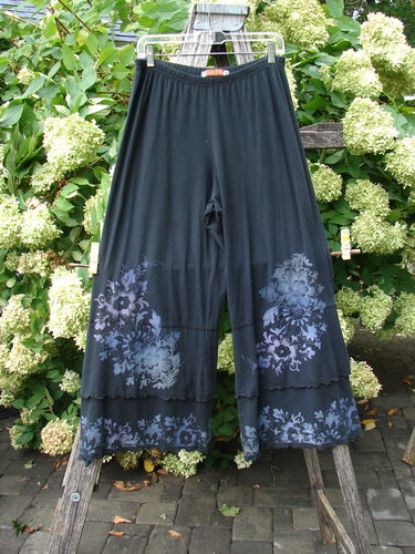 Barclay Cotton Lycra Crop Double Ruffle Pantaloon Pant Floral Black Size 1 in black cotton-lycra, with a cropped wide-leg silhouette and double ruffle hems. The sheer lower panels are printed with muted floral motifs, hanging from a clear hanger against leafy greenery.