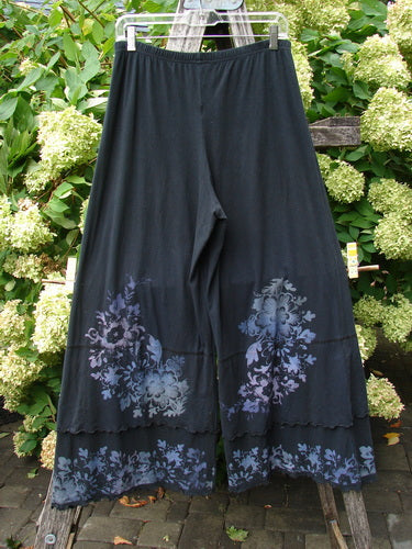 Barclay Cotton Lycra Crop Double Ruffle Pantaloon Pant Floral Black Size 1 in black cotton-lycra, with a wide cropped silhouette and double ruffle hem. Pale floral motifs decorate the lower legs, while the pant hangs centered on a hanger against dense green foliage.