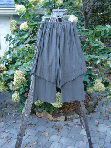 Barclay Cotton Lycra Batiste V Pocket Pant Crop Pant in Stone Grey, draped over a wooden ladder, showcasing its unique skirt-like overpiece and floppy side pocket design.