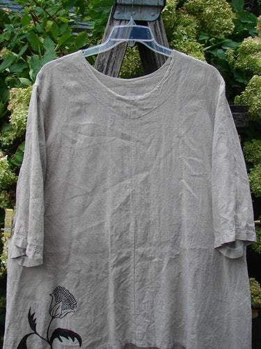Barclay Heavyweight Linen High Low Top Single Sunflower Wheat Size 2 in soft wheat linen, with a loose, draped silhouette and three-quarter sleeves. The front shows subtle wrinkling and a black sunflower motif near the hem, hanging outdoors against dense green foliage.