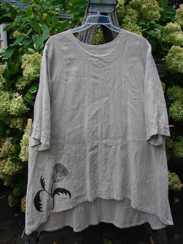 Barclay Heavyweight Linen High Low Top in wheat, size 2, with a relaxed short-sleeve silhouette, rounded neckline, and layered high-low hem. The textured linen drapes in soft folds, while a single sunflower graphic sits near the lower front in dark contrast.