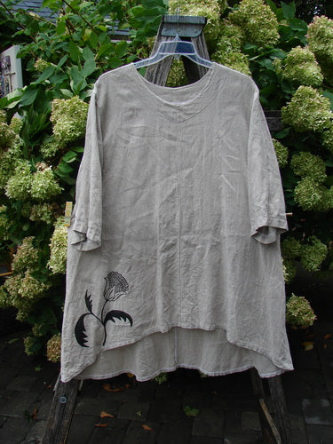 Barclay Heavyweight Linen High Low Top Single Sunflower Wheat Size 2, a loose wheat-gray linen top with a rounded neckline, draped short sleeves, and an uneven high-low hem. A black sunflower graphic sits near the lower left, and the soft, wrinkled fabric hangs against leafy greenery.