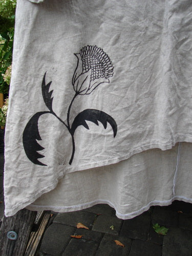 Barclay Heavyweight Linen High Low Top Single Sunflower Wheat Size 2 in wheat-toned linen, with a black single sunflower graphic and a softly frayed high-low hem. The wrinkled fabric hangs in layered folds, its matte texture and muted palette emphasized by natural outdoor light.