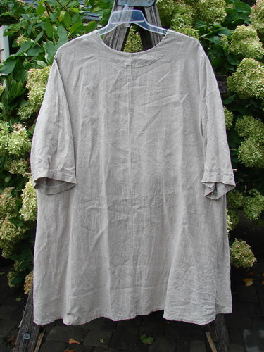 Barclay Heavyweight Linen High Low Top Single Sunflower Wheat Size 2 in soft wheat linen, shown as a loose, high-low tunic with elbow-length sleeves, a round neckline, and a gently wrinkled textured finish. It hangs on a clear hanger against dense green foliage, with muted natural light emphasizing its relaxed drape and airy silhouette.