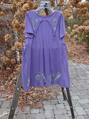 A 1994 Elfin Dress in Purple Nuit, Size 2, hangs on a rack. It features a dramatic collar, sailor-like rear collar, and a slight A-line shape. The dress has a rear tie tunnel and additional side ties for adjusting the waistline. The lower hemline widens, and there is a slight empire waist seam. The dress is made from cotton jersey and is in perfect condition.