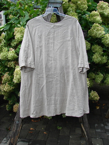 Barclay Heavyweight Linen High Low Top in wheat, size 2, hangs from a clear hanger against lush green hydrangeas. The loose linen shirt has a soft, wrinkled texture, short sleeves, and a gently curved high-low hem in a muted, natural tone.