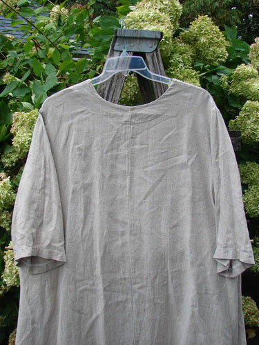 Barclay Heavyweight Linen High Low Top Single Sunflower in wheat, Size 2, shown as a loose, softly draped linen top with a rounded neckline, three-quarter sleeves, and a high-low hem. The matte fabric hangs in gentle vertical folds against a leafy garden backdrop.