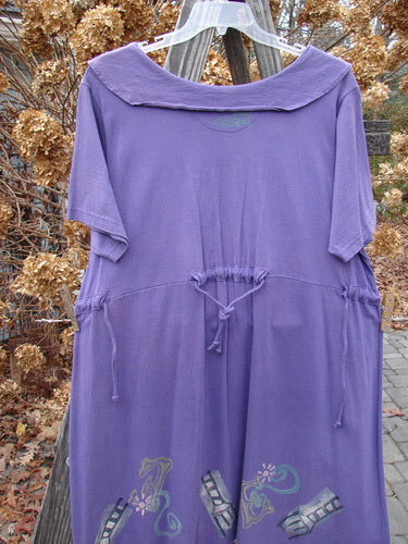 A 1994 Elfin Dress in Purple Nuit, Size 2, hangs on a clothes rack. The dress features a dramatic collar, sailor-like rear collar, and a slight A-line shape. It has a rear tie tunnel and additional side ties for adjusting the waistline. The lower hemline widens, and there is a slight empire waist seam. The dress is made from cotton jersey and is in perfect condition.