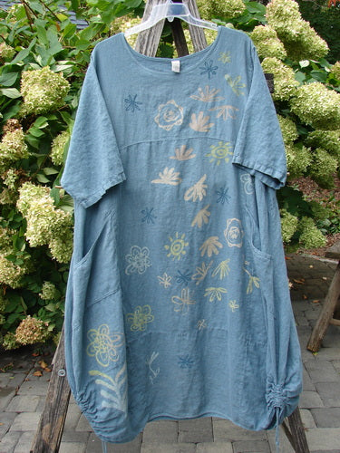 Barclay Linen Lucky Urchin Dress Floral Drift Delaware Size 2 in faded blue linen, a loose short-sleeve dress with a round neckline and scattered floral and shell-like prints in pale yellow, cream, and blue. It hangs on a wooden hanger amid lush greenery, with soft daylight bringing out the washed texture and relaxed drape.