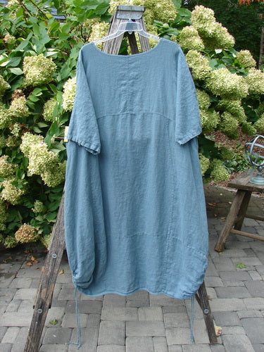 Barclay Linen Lucky Urchin Dress Floral Drift Delaware size 2 in muted blue linen, with a loose short-sleeve silhouette, softly gathered sides, and a relaxed drape. It hangs from a wooden stand outdoors, where the textured fabric contrasts with leafy greenery and gray pavers.