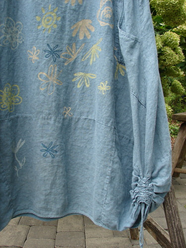 Barclay Linen Lucky Urchin Dress Floral Drift Delaware size 2 in faded blue linen, patterned with scattered floral line drawings in yellow, cream, and blue. The dress shows a relaxed drape, a curved seam, and a gathered sleeve cuff, with outdoor greenery and stone paving behind it.