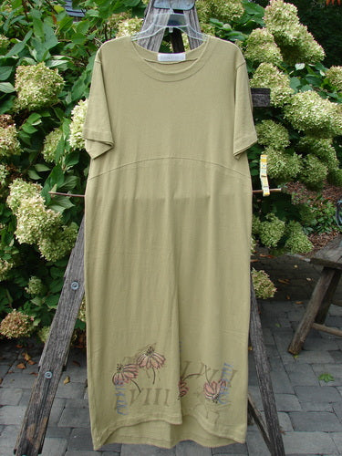 1998 Botanicals Calla Dress Echinacea Seed Size 0 in muted olive green, a short-sleeve dress with a soft crew neckline, relaxed drape, and floral graphic near the hem. The smooth fabric hangs from a wooden frame against dense green foliage.